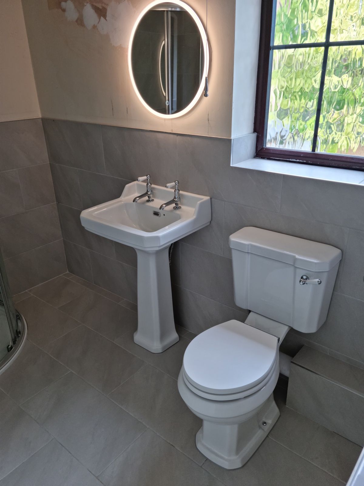 A traditional basin and toilet beneath a round lit mirror
